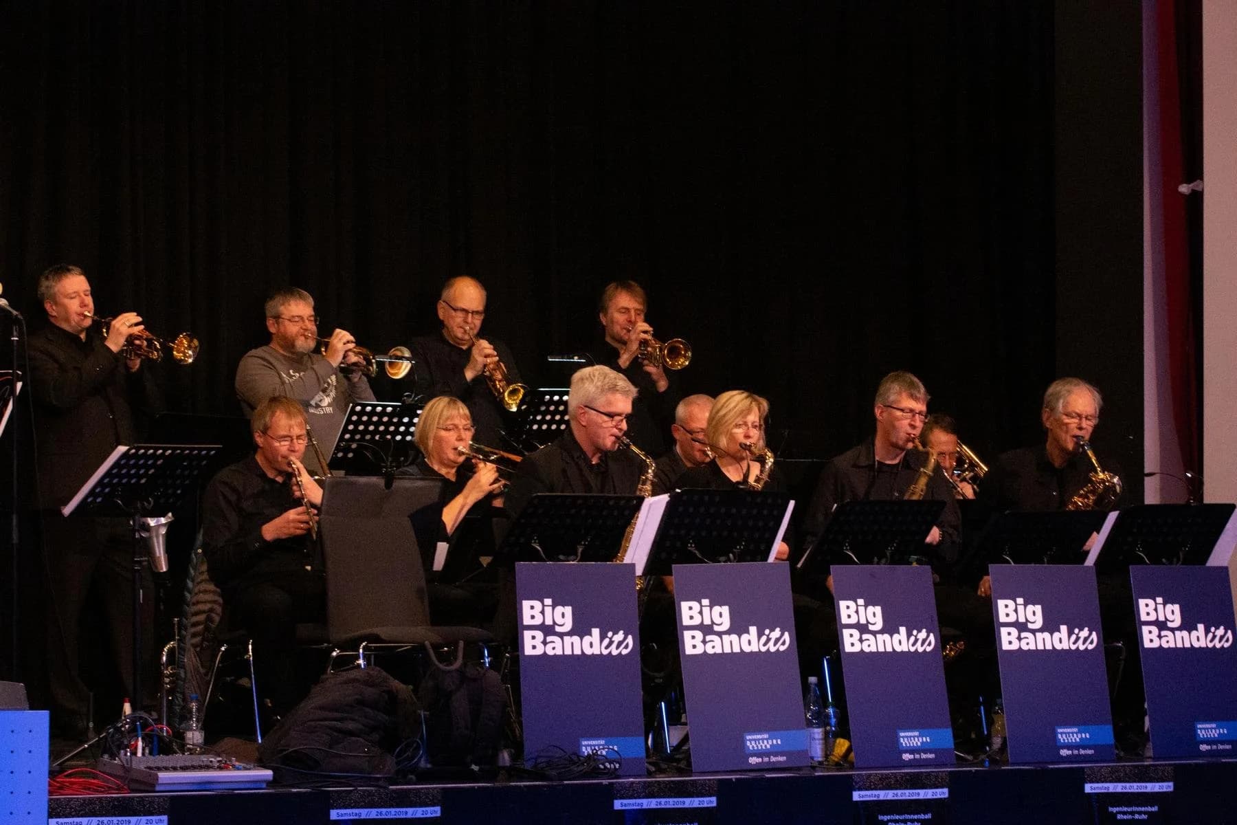 Orchester auf einer Bühne, Musiker spielen Trompeten und Saxophone unter Plakaten mit dem Text „Big Bandits“.