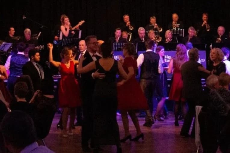 Menschen tanzen in einem Saal mit einem Live-Jazzorchester im Hintergrund.